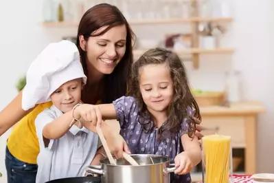 Small brother and sister cooking a meal both stirring the contents of the same pot watched over by their laughing young mother